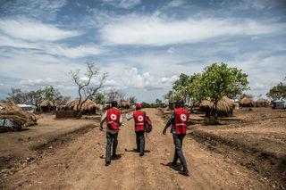 Three men walking in an open space surrounding by dirt paths and huts 